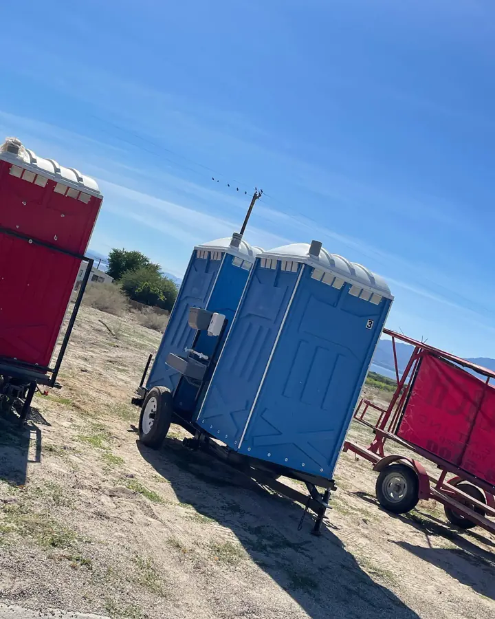 Towable trailers carrying blue and red porta potties on a dirt field