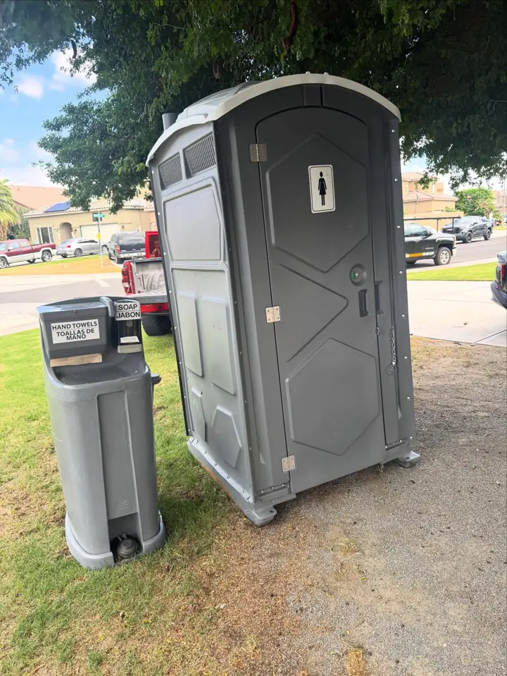 Grey standard porta potty with a freestanding handwashing station on a lawn